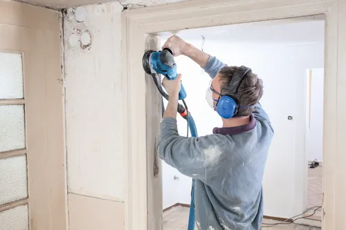 Person wearing blue ear protection, safety glasses, sanding a door frame with a blue sander, while wearing a blue sweater.