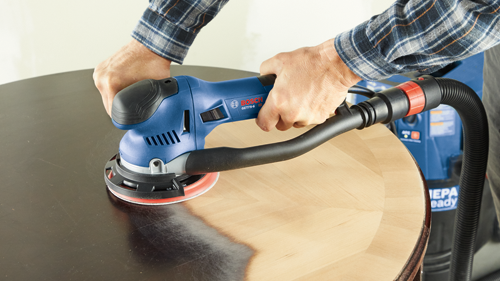 Bosch GET75-6 orbital sander being used on a round wooden surface by a person wearing a plaid shirt.