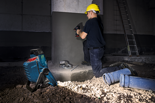 A person wearing a yellow helmet uses a drill; a Bosch work light and ladder are in the background.
