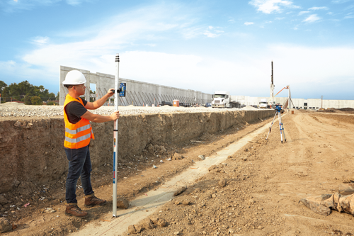 A person in a white hard hat and orange vest uses a Bosch GR 500 Professional leveling rod at a construction site.