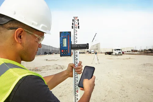 Person in construction attire, holding Bosch tool and smartphone, using a measurement rod with a visible scale.