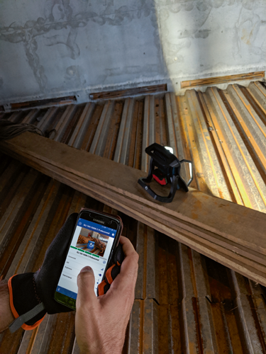 A person holding a Samsung phone displaying "GLI 18V-10000 C Professional" on its screen, using the app to adjust the brightness of a work light. A Bosch GLI 18V-10000 C Professional work light is also visible.