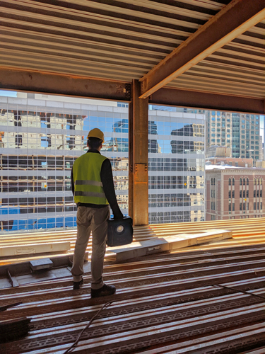 A person wearing a yellow helmet and vest looks out a window at a city skyline, holding a black rectangular object in their left hand.