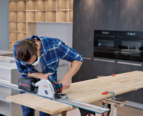 A person wearing a blue plaid shirt, safety glasses, and a face mask uses a Bosch track saw to cut a wooden plank.
