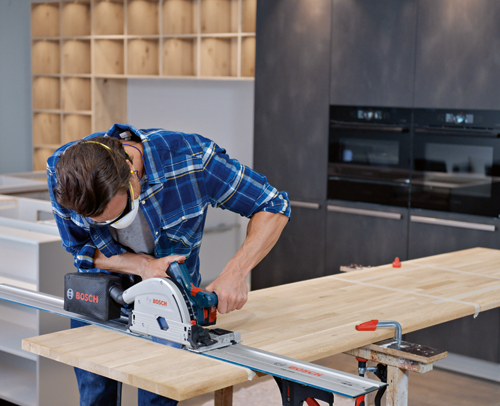 A person wearing a blue plaid shirt, safety glasses, and a face mask uses a Bosch track saw to cut a wooden plank.