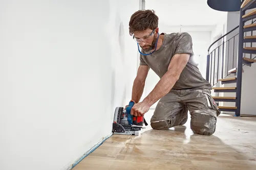 A person wearing safety glasses kneels to operate a circular saw near a wall.
