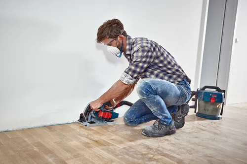 A person wearing safety glasses and a mask cuts a wooden floor with a Bosch circular saw. Bosch Professional vacuum cleaner nearby.