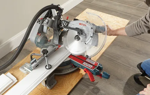Bosch miter saw cutting trim, with a person holding the saw and wearing a knee pad.