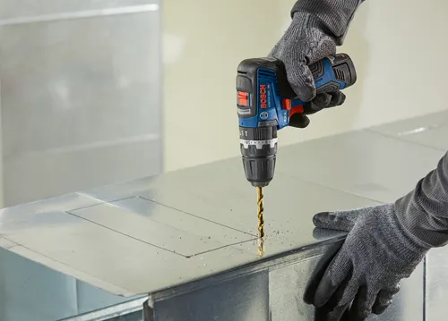 Bosch GSB12V-300 HD cordless drill being used to drill a hole in a metal sheet. The user wears grey gloves. Visible text: "BOSCH" and "GSB12V-300 HD".