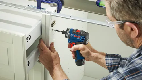 Person wearing glasses and a plaid shirt using a Bosch GSR12V-300HX HD drill to attach a cabinet drawer.