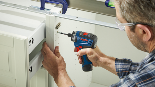 Person wearing glasses and a plaid shirt using a Bosch GSR12V-300HX HD drill to attach a cabinet drawer.