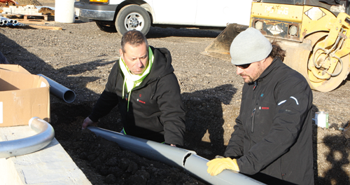 Two people install a pipe. One wears a black jacket with the Bosch logo and yellow gloves. The other wears a black jacket with the Bosch logo.