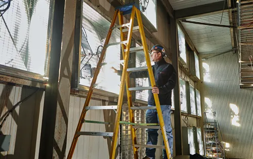 Person wearing glasses and a hard hat stands on a yellow ladder, holding onto it in a metal building, wearing a Bosch jacket.
