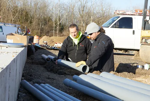 Two men in work clothes examining gray pipes, one with a Bosch logo on the jacket.