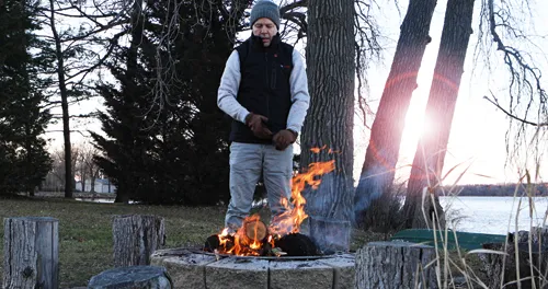 A person stands near a campfire, wearing a hat, gloves, and vest, with trees, grass, and a body of water in the background.