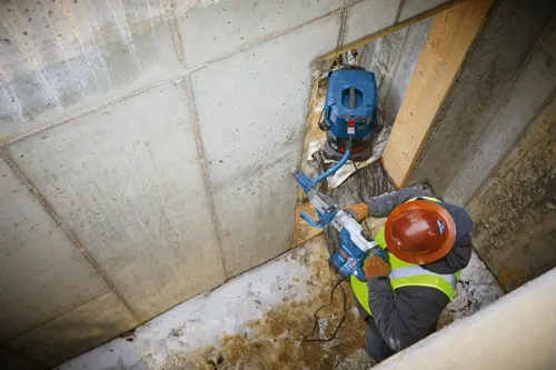 Person wearing a hard hat and safety vest uses a Bosch rotary hammer, while a Bosch HEPA READY vacuum is nearby.
