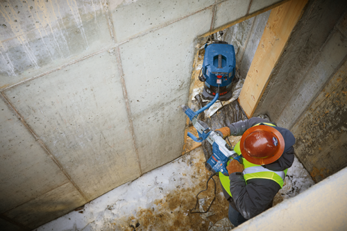 Person wearing a hard hat and safety vest uses a Bosch rotary hammer, while a Bosch HEPA READY vacuum is nearby.
