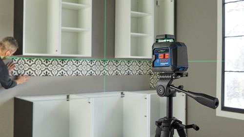 Bosch Professional GLL50-40G green laser level on tripod, emitting green laser beams, with a person placing decorative tiles.