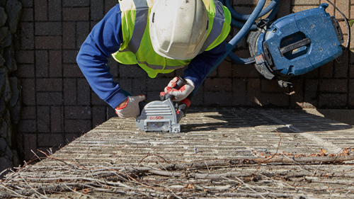 A person wearing a white hard hat and safety vest uses a Bosch cutting tool labeled 'BOSCH' on a brick surface; a blue vacuum cleaner is also visible.