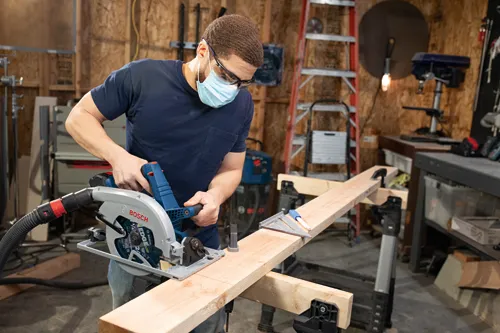 Person wearing glasses and mask, using a Bosch circular saw on a wooden plank in a workshop.