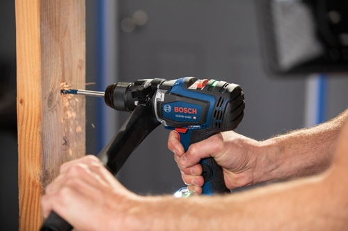 Bosch GSB18V-1330C drill being used to drill into a wooden board. The drill is blue and black, the board is light brown, and sawdust is flying.