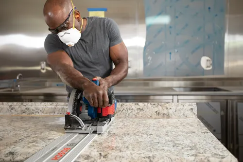 A person of African descent wearing safety glasses and a mask using a Bosch track saw to cut a countertop. The track saw is on a guide rail with "BOSCH" visible.