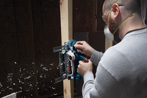 A person wearing safety glasses and a mask uses a Bosch PLH32 power planer to work on a wooden frame.