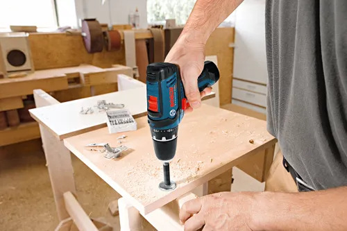 Bosch Lithium-Ion 12V cordless drill being used by a person to drill a hole in a wooden board. The person is wearing a gray shirt.
