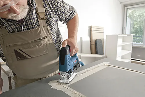 A person wearing overalls using a blue and black jigsaw to cut a grey surface.