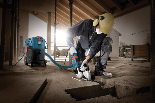 A person wearing safety gear, cuts into concrete with a Bosch saw, with a vacuum cleaner nearby to collect debris.