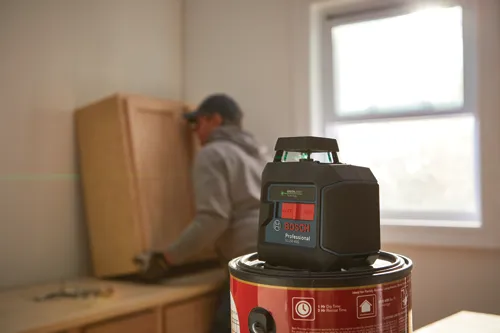 Bosch Professional GLL50-40G level, with OFF and ON buttons, on a can of paint, and a person installing a cabinet in the background.