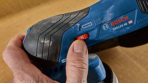 Bosch GSS12V-40 HD cordless sander, a hand presses the red "6" setting on the speed adjustment dial.