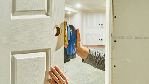 Bosch GSS12V-40 sander is being used to sand a door; the sander has a yellow sanding surface, and the hand of a person is visible holding the sander.