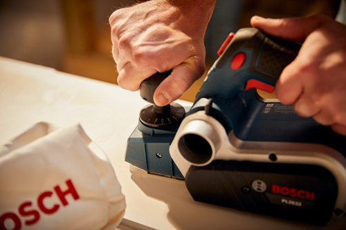 Bosch PL2632 power planer with visible depth markings, held by a person. A dust collection bag with the  word "OSCH" is visible.