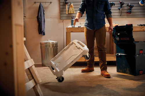 Person in brown pants and denim shirt next to a stack of Bosch L-Boxx cases, and rolling a white L-Boxx. Other tools and a trash can are also in the garage.