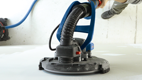 A person holds a blue and black circular sanding tool with a flexible hose. The tool is on a white surface.