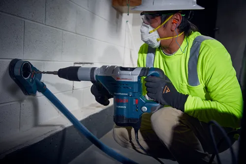 Person wearing hard hat, safety glasses, and mask operating a Boschhammer BOSCH 11264EVS, drilling into a concrete block wall.