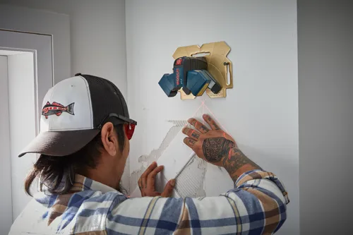 A person installs a tile, using a Bosch GTL3 laser level. The laser level is mounted on a wall and projects red lines. The person wears a plaid shirt and a cap with a fish graphic.