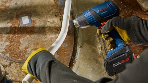Person wearing work gloves using a Bosch GHG18V-50 heat gun to bend a white plastic pipe. The heat gun is powered by a CORE18V 8 Ah battery.