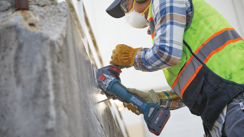 Person using a Bosch GWX18V-10P angle grinder, wearing safety glasses, a respirator mask, gloves, a reflective vest, and a cap.