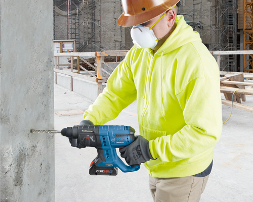 Person wearing a hard hat, face mask, and high-visibility jacket operating a Bosch GBH18V-21 hammer drill to drill into a concrete column.