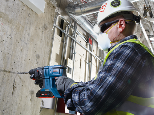 Bosch GBH18V-21 hammer drill being used by a person wearing safety glasses, ear plugs, a face mask, and a hard hat.