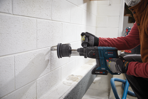 Bosch GBH18V-21 drill being used to drill into a white cinder block wall by a person wearing gloves and a respirator mask.
