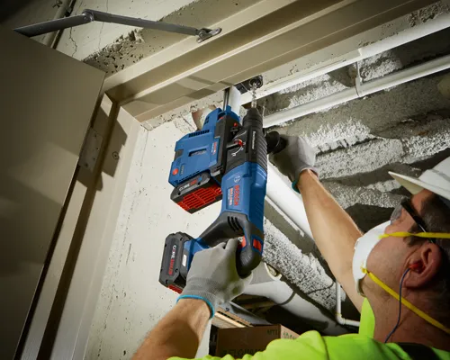 Bosch BULLDOG GBH18V-26D and GDE18V-26D drills, a worker wearing safety glasses, a face mask, and earplugs drilling into a ceiling.