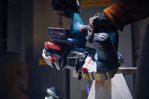 Bosch GCB18V-2 band saw in use, cutting a piece of wood, visible 18V battery. The tool has red and white accents, and is being held by a gloved hand.