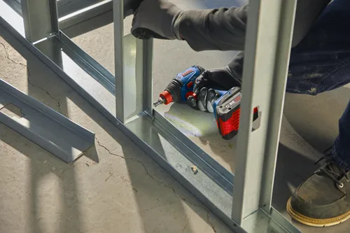 Person wearing gloves using a Bosch GDX18V-1800C CORE18V impact driver to work on a metal stud wall.