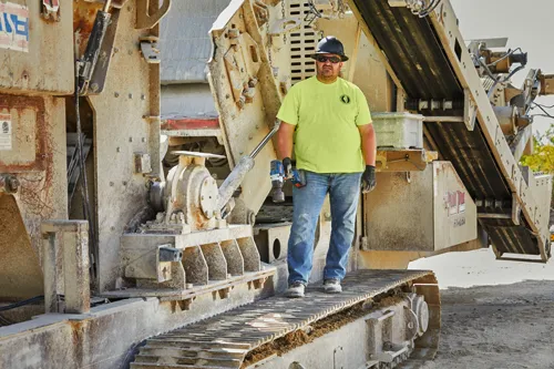 A person wearing a black hard hat, sunglasses, a yellow shirt, blue jeans, and black gloves stands on the tracks of a large machine, holding a Bosch cordless impact wrench.