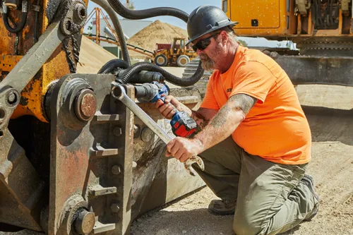 A person wearing a hard hat, sunglasses, and an orange shirt, is kneeling while using a wrench and a power tool to work on construction machinery.