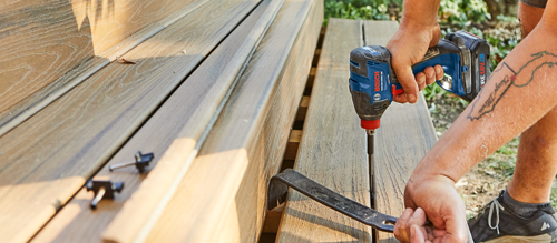 Bosch cordless impact driver, a crowbar, and screws are used to assemble a wooden bench; a person with a tattoo on their arm.