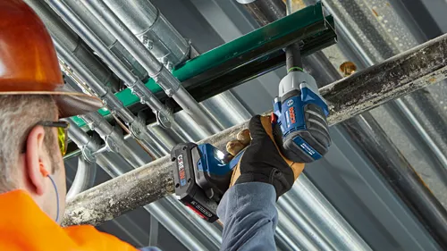 Person wearing orange hard hat and safety glasses using a Bosch cordless impact wrench, with visible 'Bosch' and 'BRUSHLESS MOTOR' text, installing above pipes.